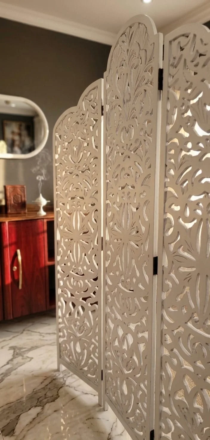 A white carved three panel folding screen standing on marble beside a red cabinet